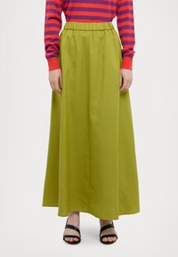 Gonna maxi verde con elastico in vita, realizzata in un tessuto leggero e liscio. Abbinata a sandali neri con cinturini e a una maglietta a righe rossa e viola.
