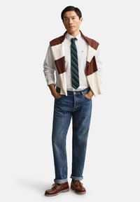 Mand iført hvid skjorte med knapper, grønstribet slips, blå jeans, brun- og hvidstribet sweater hængende over skuldrene og brune sko.