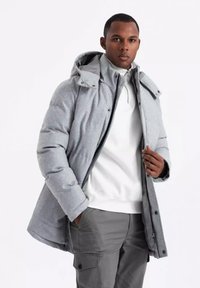 Abrigo acolchado gris con capucha, con cremallera frontal y botones a presión, llevado sobre un suéter blanco, combinado con pantalones grises.