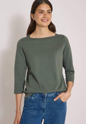 Femme souriante, portant un haut vert à manches longues et un jean bleu, une main dans la poche sur un fond uni.