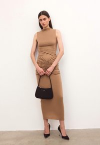 Naine beezhise ilma varrukateta ruched midi dresses, käes väike must käekott, jalas mustad kõrged kontsad, seismas lihtsa valge seina ees.