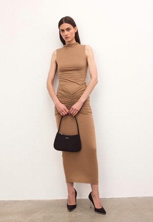 Eine Frau in einem ärmellosen, sandfarbenen Midikleid mit Raffung, die eine kleine schwarze Handtasche hält und schwarze High Heels trägt, steht vor einer schlichten weißen Wand.
