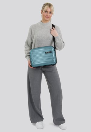 Vrouw in een grijze trui en broek die een teal hard-shell schoudertas met zwarte riem vasthoudt, staand tegen een effen lichte achtergrond.