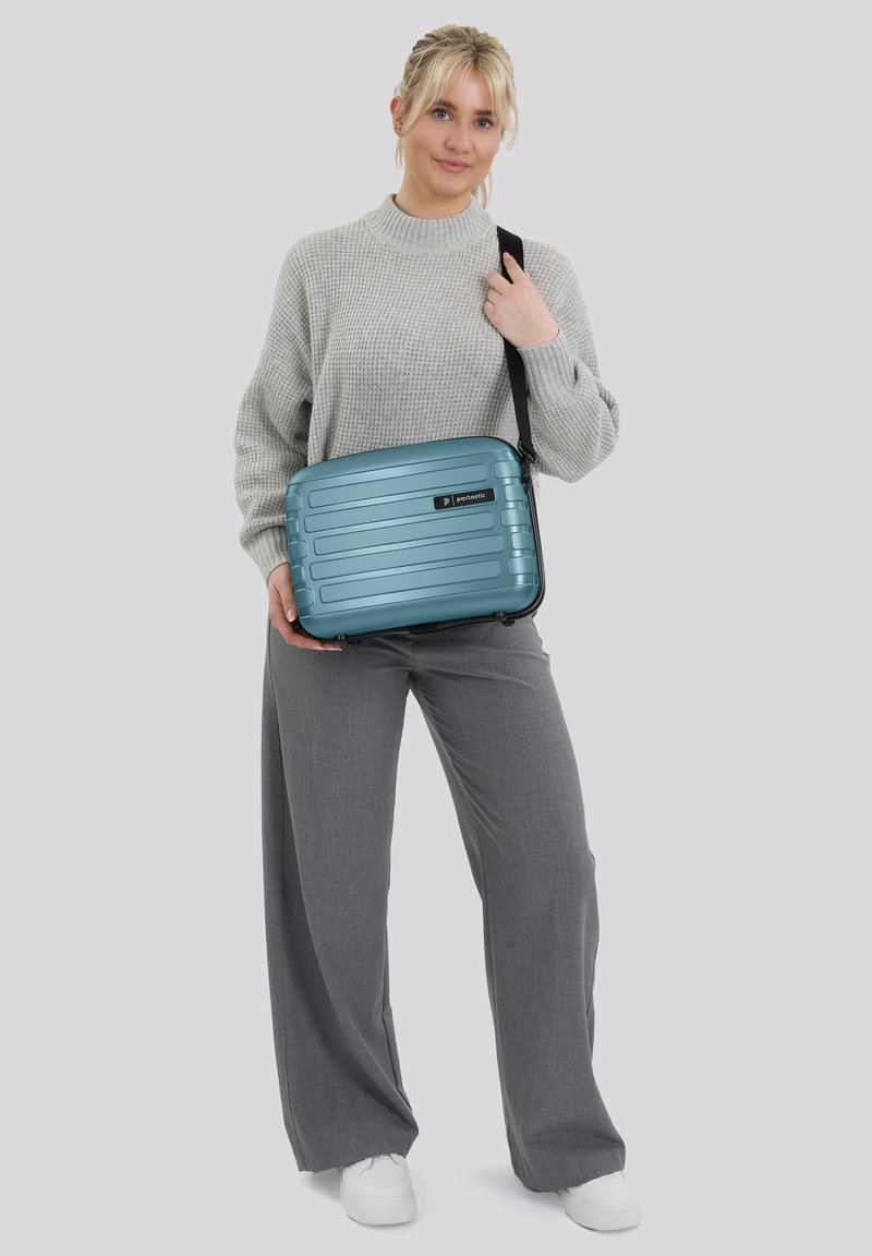 Donna in maglione grigio e pantaloni che tiene una borsa a tracolla rigida color turchese con tracolla nera, in piedi contro uno sfondo chiaro e uniforme.