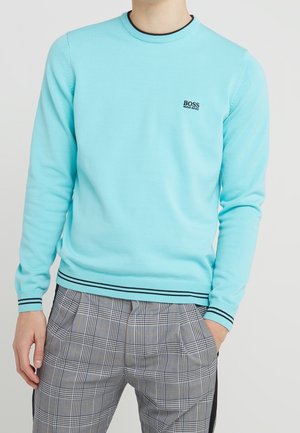 Homme portant un pull Hugo Boss bleu clair avec des bordures noires et un pantalon à carreaux gris, debout avec une main dans la poche.