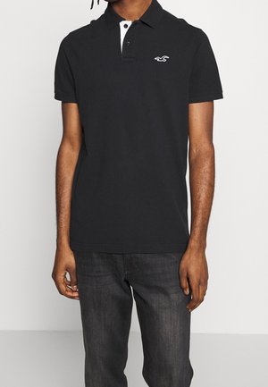 Hombre con camisa tipo polo negra de manga corta con botonadura blanca y un pequeño logo blanco en el pecho, combinado con jeans oscuros.