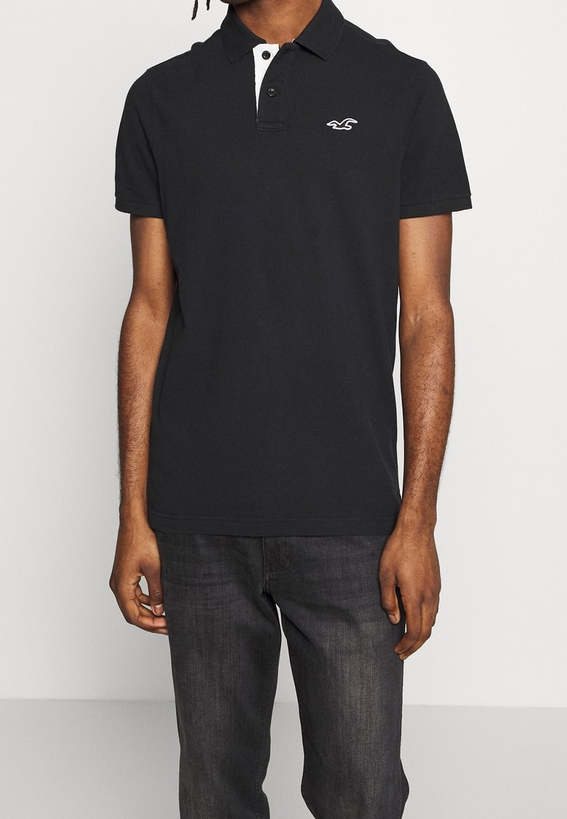Homme portant un polo noir à manches courtes avec une patte de boutonnage blanche et un petit logo blanc sur la poitrine, assorti à un jean foncé.