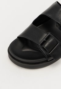 Sort lædersandal med åben tå, to brede stropper og en rektangulær spænde. Struktureret overflade og en rund sort gummisål.
