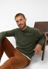Dunkelgrüner Strickpullover mit strukturiertem Muster, kombiniert mit braunen Cordhosen mit vertikalen Streifen, neben einem Holztstuhl sitzend.