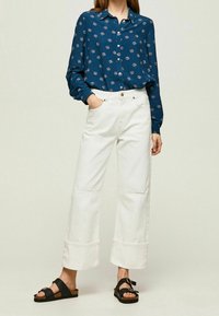 Blå skjorta med knappar och blommönster, vita vida byxor med kontrastsömmar och svarta sandaler med remmar och spänne.