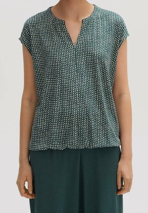 Frau trägt eine grüne kurzärmelige Bluse mit weißem kleinem abstraktem Punktmuster und passenden dunkelgrünen Hosen.