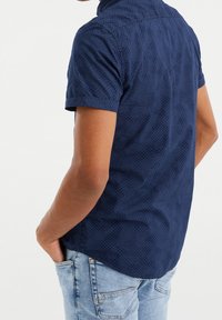 Námořnicky modrá košile s krátkým rukávem a jemným geometrickým vzorem, se zapínáním na knoflíky u límce a zaobleným lemem, zastrčená do světle modrých džín.