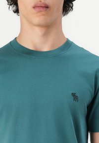 Türkises Baumwoll-T-Shirt mit Rundhalsausschnitt und kurzen Ärmeln, versehen mit einem dezenten gestickten Design eines Hirsches im linken Brustbereich.