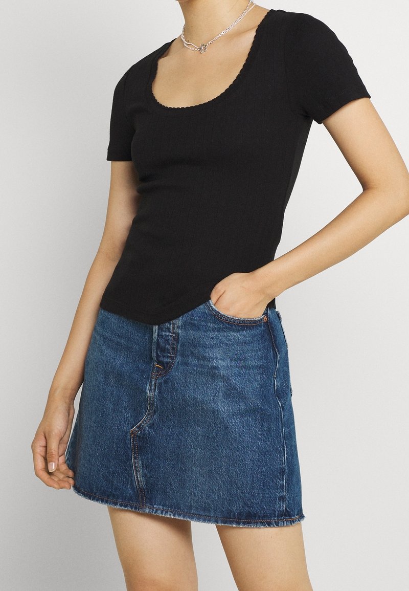 Personne portant un haut côtelé noir à manches courtes avec un col rond et une mini-jupe en denim bleu, avec une couture à l'avant et une fente.