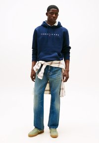 Homem jovem vestindo capuz azul marinho com o logótipo "Tommy Jeans", jeans azuis, camisa bege xadrez amarrada à cintura e ténis verdes sobre fundo branco.