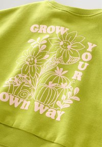 Lichtgroene trui met glimlachende bloemen, planten, een slak en de zin "Grow your own way" in witte tekst en illustraties.