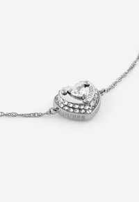 Srebrna bransoletka w kształcie serca z widocznym kryształem na środku, ozdobiona mniejszymi kamieniami i grawerowanym logo z boku.