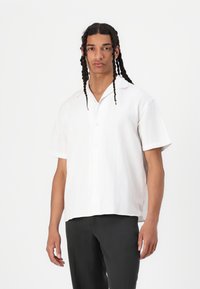 Short-sleeve white linen shirt with a collared neckline, button front, and a relaxed fit, paired with dark trousers.