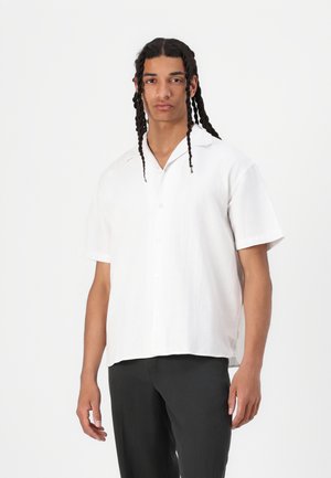 Chemise en lin blanche à manches courtes avec un col, des boutons à l'avant et une coupe décontractée, assortie à un pantalon sombre.