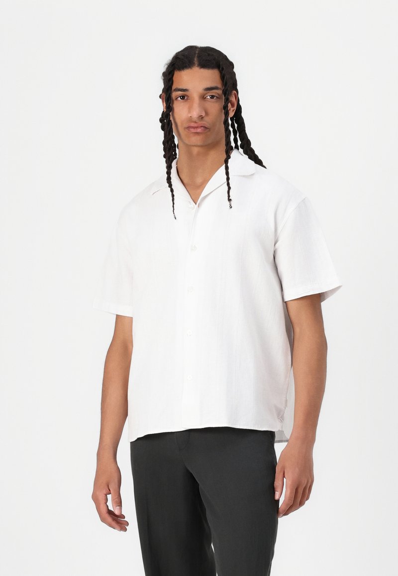 Short-sleeve white linen shirt with a collared neckline, button front, and a relaxed fit, paired with dark trousers.