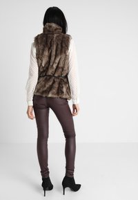 Braune Kunstpelzweste mit hohem Kragen, tailliertem Gürtel, geschichtete Textur; kombiniert mit eng anliegenden braunen Hosen und schwarzen Stilettostiefeln.