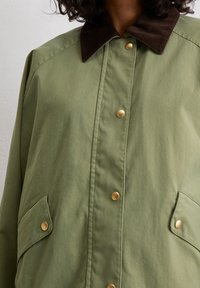 Olive green jacket with a brown corduroy collar. Features gold snap buttons and two side pockets. Smooth fabric with a relaxed fit.