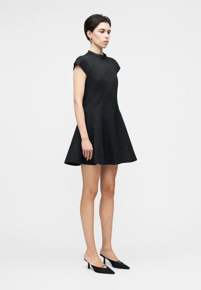 Juoda rankovių neturinti suknelė su aukštu kaklu, išplatėjusia suknelės dalimi ir matomomis siūlių detalėmis; derinama su smailiais juodais aukštakulniais ant neutralios spalvos fono.