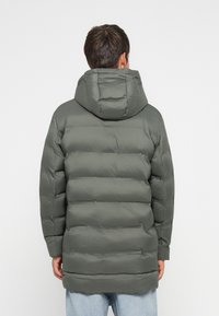 Παλτό με γέμισμα σε σκούρο πράσινο χρώμα με κουκούλα, διαθέτοντας ραφές τύπου κουβέρτα, μακριά μανίκια και μεσαίου μήκους κόψιμο, φαίνεται από πίσω.