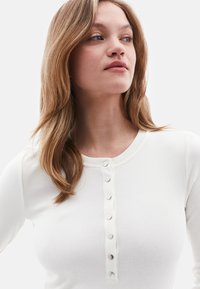 Witte geribbelde longsleeve top met een ronde halslijn en een stolpplacket met vijf metalen knoopaccenten aan de voorkant.