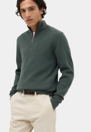 Junger Mann trägt einen dunkelgrünen gerippten Strickpullover mit Viertel-Reißverschluss und hellbeige Hose, Hände in den Taschen, vor einem einfarbigen Hintergrund.