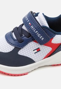 Sneaker bianco in rete con accenti blu navy e rossi. Presenta una fascia regolabile, suola testurizzata e marchio sulla fascia e sul lato.