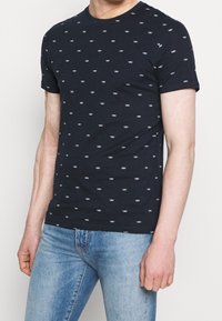 Navyblaue Baumwoll-T-Shirt mit kurzen Ärmeln, das ein wiederholtes kleines weißes Bootmuster zeigt, klassischen Rundhalsausschnitt und figurnahe Passform.