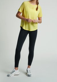 Kobieta w żółtej koszulce z krótkim rękawem, czarnych legginsach i białoszarych butach sportowych stoi na jednolitym tle.