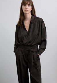 Donkerbruine, gestructureerde outfit met een wikkelstijl top met kraag en elegante geplooide broek, beide met een subtiel patroon.