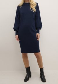 Abito in maglia blu navy con maniche lunghe a palloncino, scollo rotondo e una vestibilità comoda, abbinato a stivali neri alla caviglia.