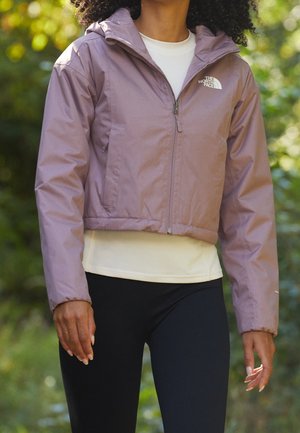 Veste courte violet clair avec capuche, fermeture éclair et poches latérales. Associée à une chemise ajustée crème et à des leggings noirs.