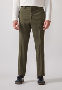 Olivgrüne Hose mit geradem Schnitt, die Falten vorne aufweist, aus glattem Material, kombiniert mit schwarzen Sportschuhen.