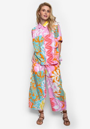 Vrouw staand, gekleed in een kleurrijk loszittend shirt en broek met roze, oranje, blauwe en witte abstracte bloemenpatronen, en bruine sandalen met bandjes.