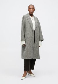 Manteau en chevrons gris avec larges revers, manches surdimensionnées, porté sur un haut texturé blanc et un pantalon foncé. Des ballerines blanches pointues complètent la tenue.