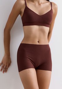 Vrouw in een bijpassende donkerbruine naadloze beha en boyshort onderbroek, staand tegen een lichtgekleurde muur met één hand erop leunend.
