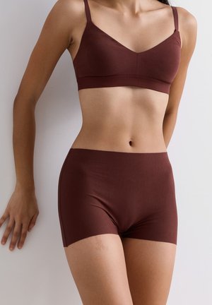 Frau in passendem, dunkelbraunem nahtlosem BH und Boyshort-Unterwäsche, die an einer hellen Wand steht und eine Hand darauf resting.