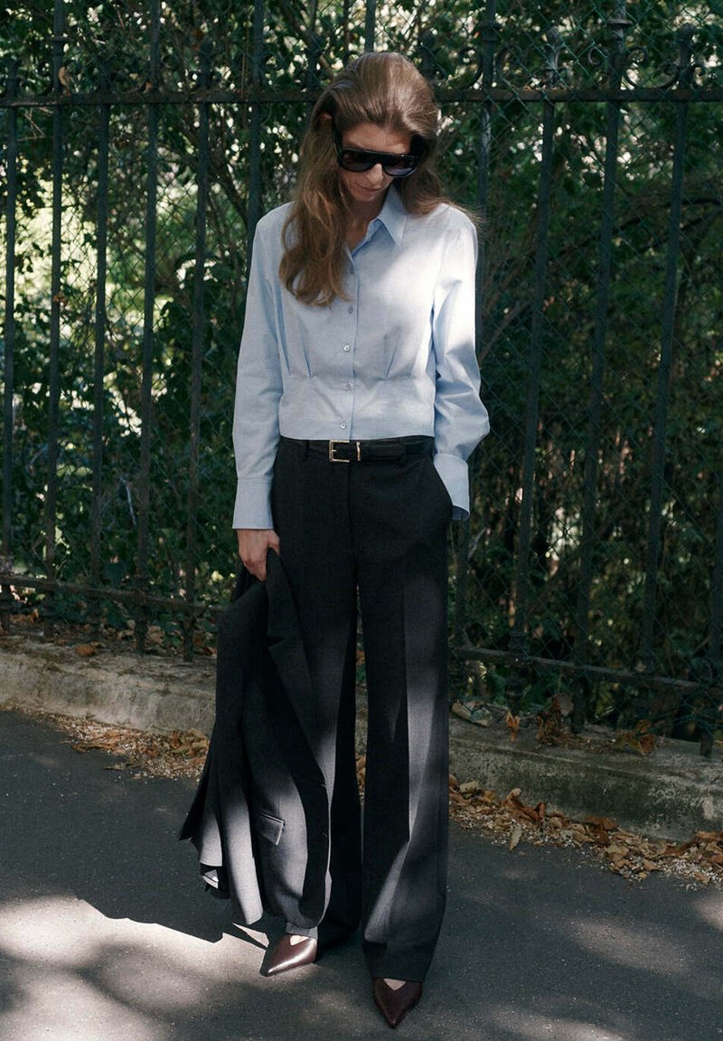 Femme portant des lunettes de soleil, chemise bleu clair boutonnée, pantalon large noir, tenant une veste noire, debout à l'extérieur près d'une clôture avec de la verdure.