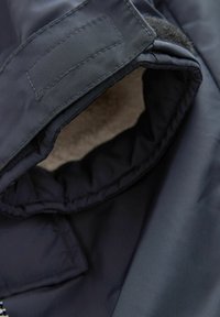 Jachetă bleu marin cu un exterior texturat, rezistent la apă. Interiorul are un căptușeală moale. Include închidere cu Velcro pe mânecă.