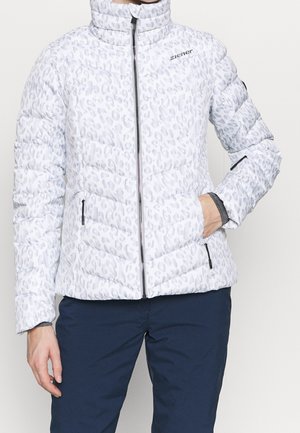 Veste matelassée blanche avec imprimé léopard gris clair, fermeture éclair à l'avant, poches zippées, et pantalon bleu marine, une main dans la poche.
