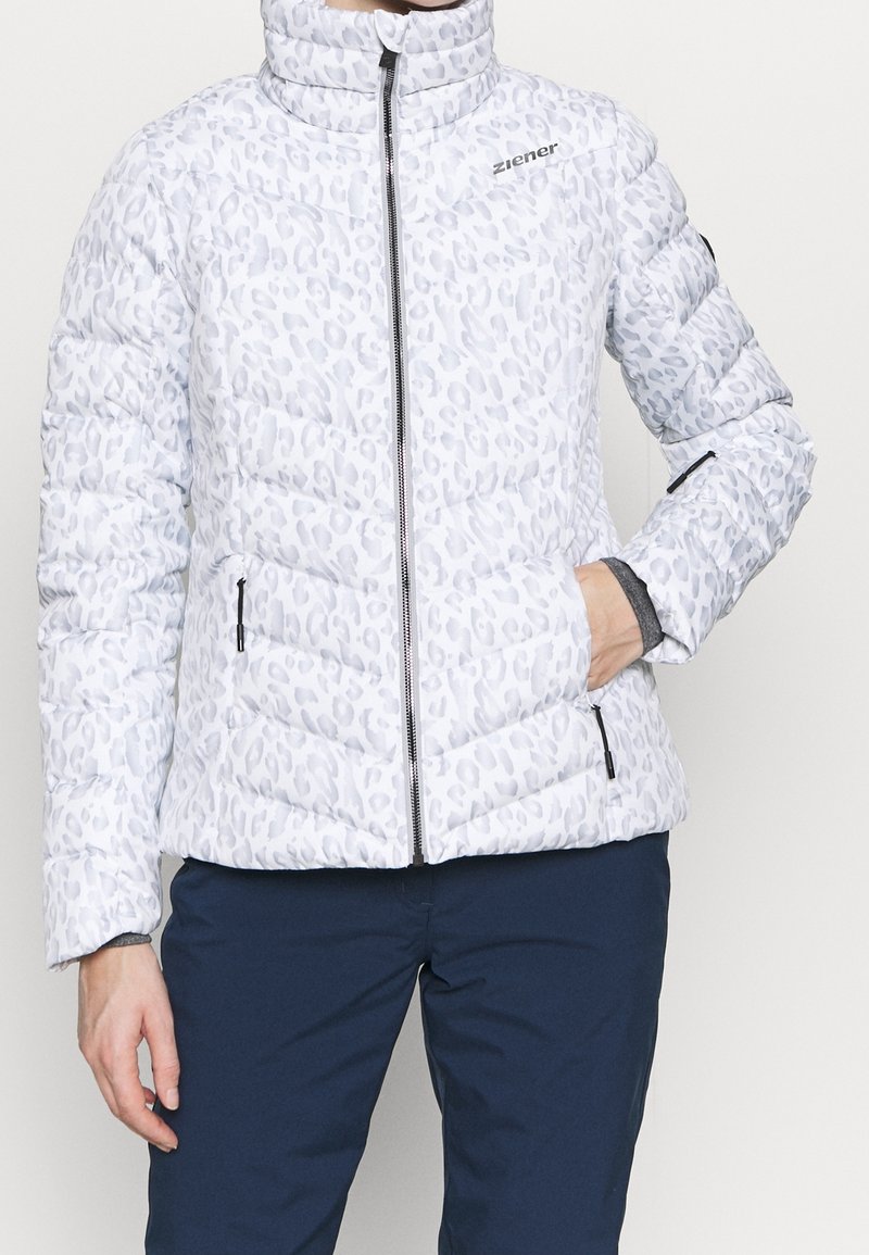 Veste matelassée blanche avec imprimé léopard gris clair, fermeture éclair à l'avant, poches zippées, et pantalon bleu marine, une main dans la poche.