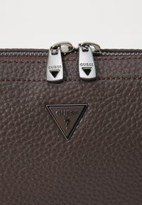 Bolso de cuero marrón texturizado con un cierre de cremallera plateado, que presenta dos tiradores metálicos y un acento de logotipo metálico triangular en la parte frontal.