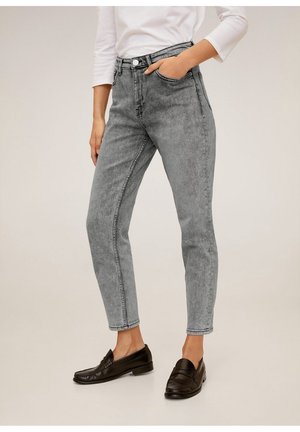 Persoon die een hoge taille grijze spijkerbroek met zuurwasteffect draagt, gecombineerd met zwarte loafers en een wit shirt met lange mouwen, met één hand in de zak.