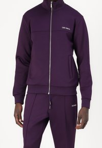 Chándal morado con chaqueta con cremallera y cuello alto, bolsillo delantero y logo bordado. Tejido liso con puños y cinturilla ajustados.