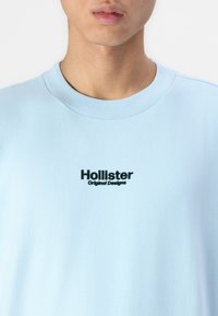 Lys blå crew neck t-skjorte laget av mykt stoff, med en svart "Hollister"-logo med "Original Designs" under. Enkel, uformell design.