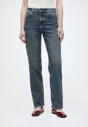 Person iført højtaljede, lige blå jeans og røde lakballetsko, stående på en hvid baggrund.